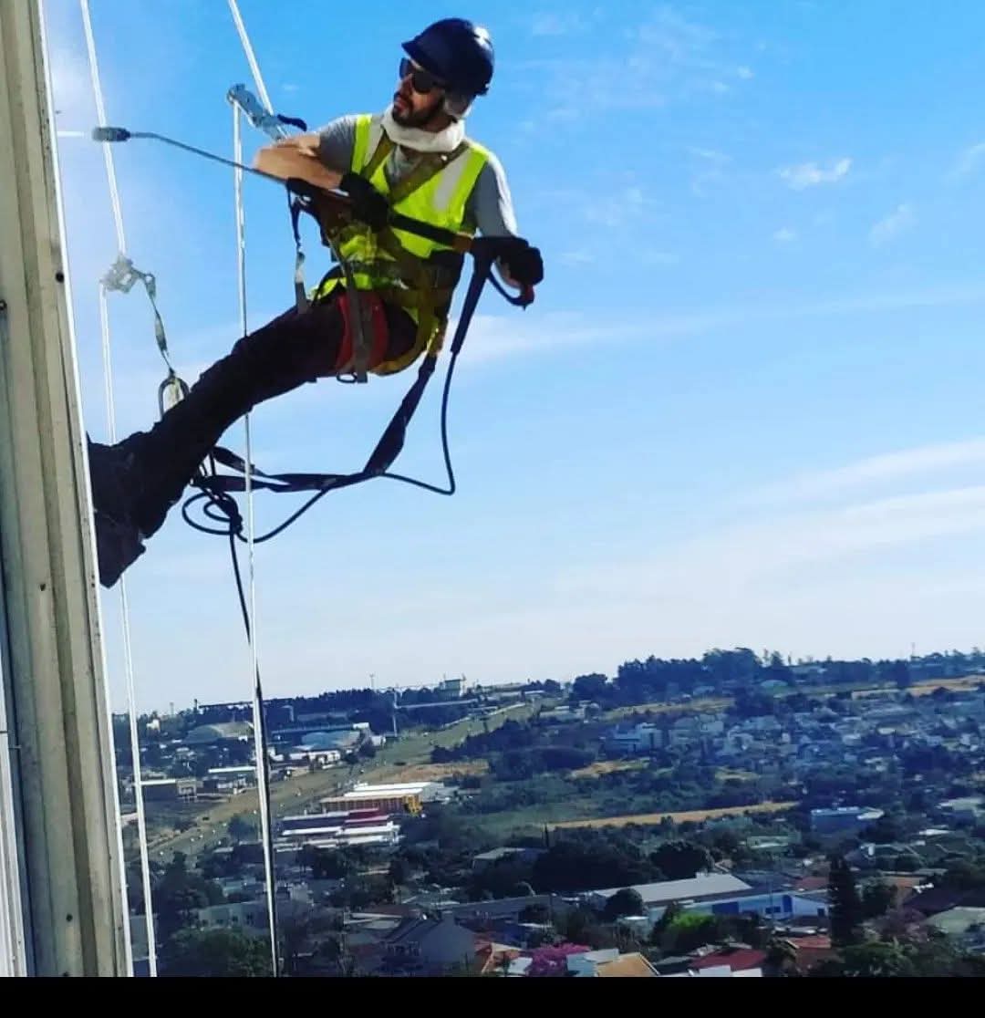 Aerial view of a cleaning crew in a modern office space, with two individuals using mops and a vacuum cleaner, and one person cleaning stairs. All are dressed in blue overalls and wearing yellow gloves. The environment features plants and contemporary furniture.